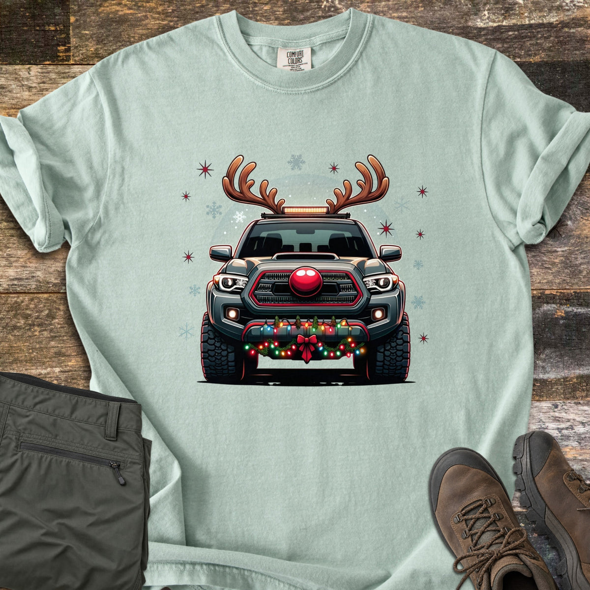 Santa Truck Comfort Colors T-shirt in Bay, laid flat. A gray pickup truck wearing reindeer antlers and a red nose, with string lights and a red bow on the bumper, among stars and snowflakes. Laid on a wood floor with olive pants and brown hiking boots.