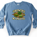 Growing My Palette Comfort Colors crewneck sweatshirt in Blue Jean, laid flat. A wooden artist's palette covered in mounds of green moss, with a moss-tipped paintbrush, sits between the moss-draped words Growing My Palette. Laid flat on a white background.