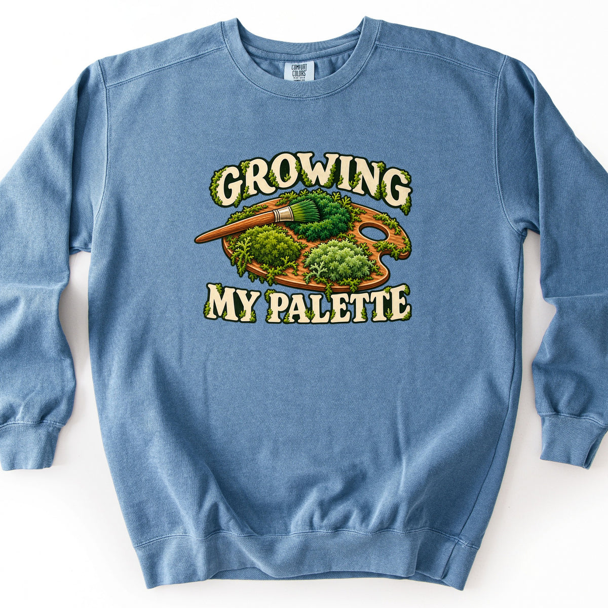 Growing My Palette Comfort Colors crewneck sweatshirt in Blue Jean, laid flat. A wooden artist's palette covered in mounds of green moss, with a moss-tipped paintbrush, sits between the moss-draped words Growing My Palette. Laid flat on a white background.