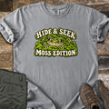 Hide & Seek Moss Edition Comfort Colors T-shirt in Grey, laid flat. A wide-eyed frog peeks out from a mound of green moss between the words Hide & Seek and Moss Edition. Olive cargo pants and brown hiking boots lie beside it on weathered wood planks.