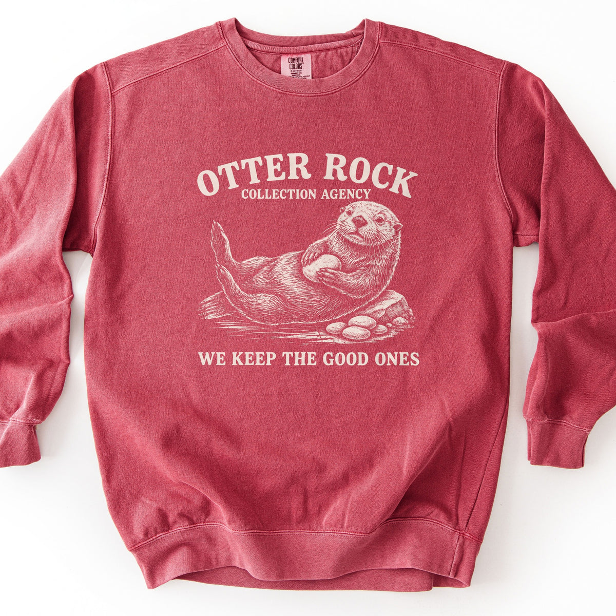 Otter Rock Collection Agency Comfort Colors crewneck sweatshirt in Crimson, laid flat. A cream sketch-style sea otter floats on its back clutching a round stone, beside a rock and a pile of pebbles, between the words Otter Rock Collection Agency and We Keep the Good Ones. Shown on a white background.