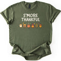 Smore Thankful Comfort Colors T-shirt in Military Green, laid flat. Cream letters read S'MORE THANKFUL above a row of smiling cartoon characters: a marshmallow, a s'more, a cocoa mug, a campfire, a pinecone and an acorn among autumn leaves. Sleeves rolled, on white.