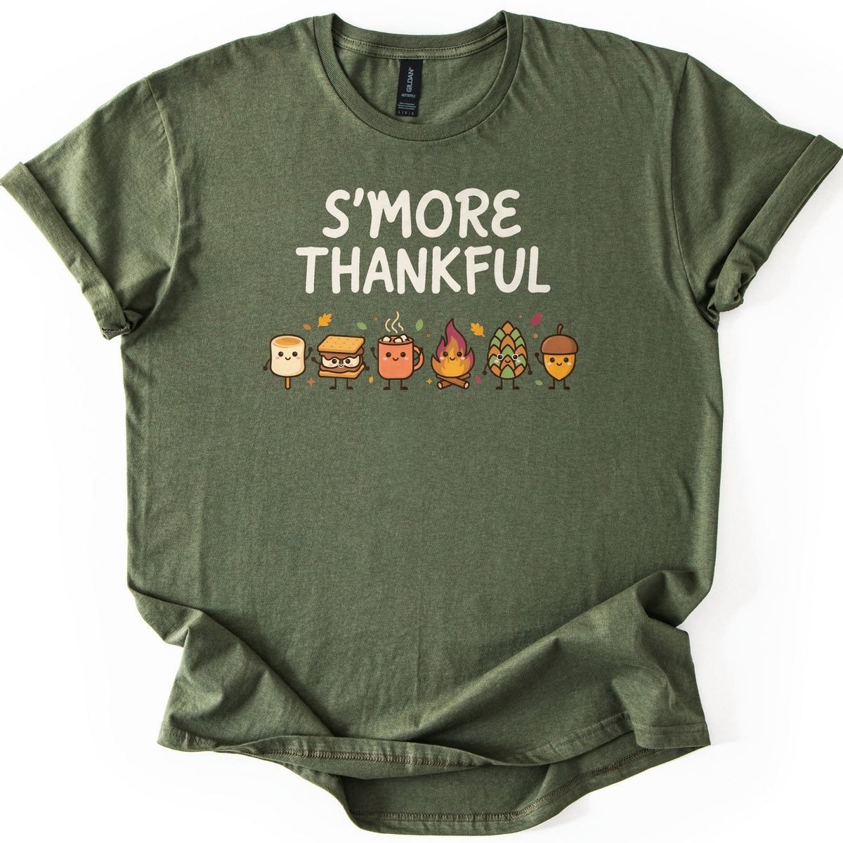 Smore Thankful Comfort Colors T-shirt in Military Green, laid flat. Cream letters read S'MORE THANKFUL above a row of smiling cartoon characters: a marshmallow, a s'more, a cocoa mug, a campfire, a pinecone and an acorn among autumn leaves. Sleeves rolled, on white.