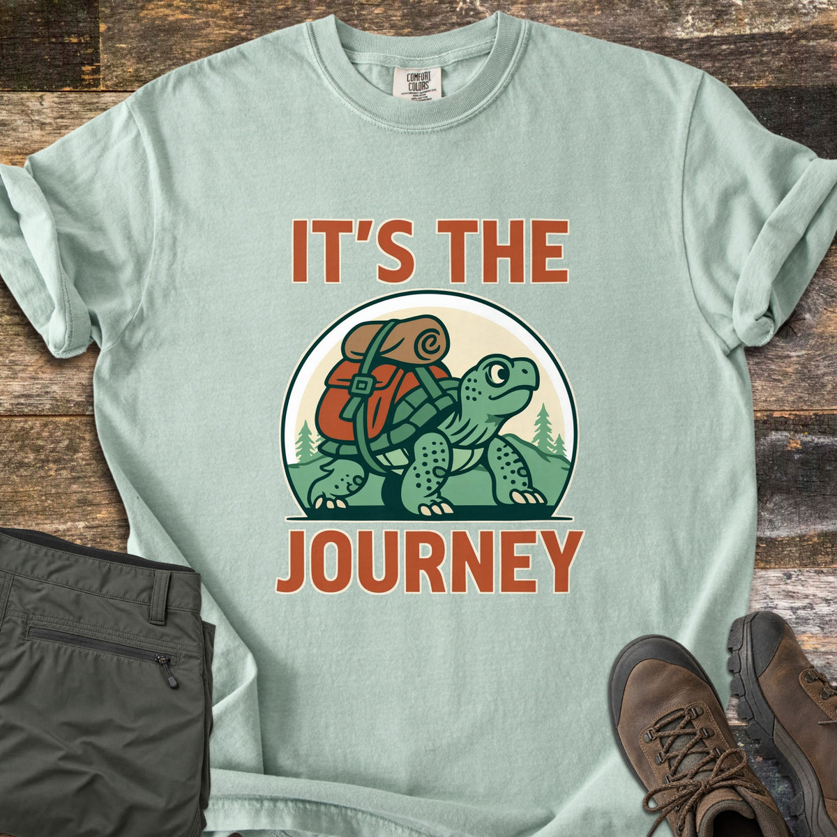 Slow Turtle Hiker Journey Comfort Colors T-shirt in Bay, laid flat. A cartoon turtle carrying a red backpack and bedroll walks past pine trees and hills between the words It's the Journey. Olive hiking pants and brown hiking boots sit beside it on rustic wood planks.
