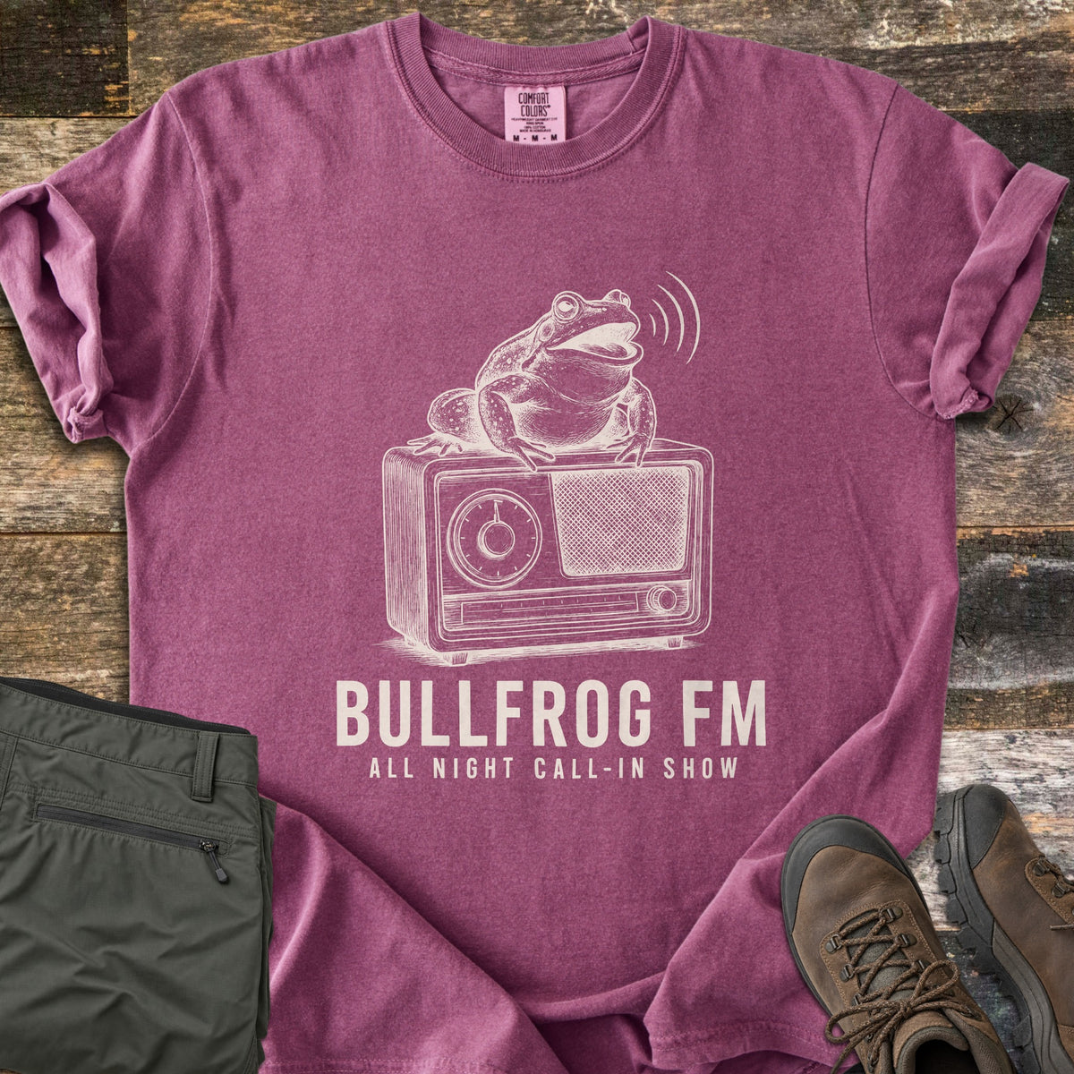 Bullfrog FM Comfort Colors T-shirt in Berry, laid flat. Cream illustration of a bullfrog croaking on a vintage tabletop radio, with sound waves, above the words BULLFROG FM and ALL NIGHT CALL-IN SHOW. Sleeves rolled on rustic wood beside olive pants and brown hiking boots.
