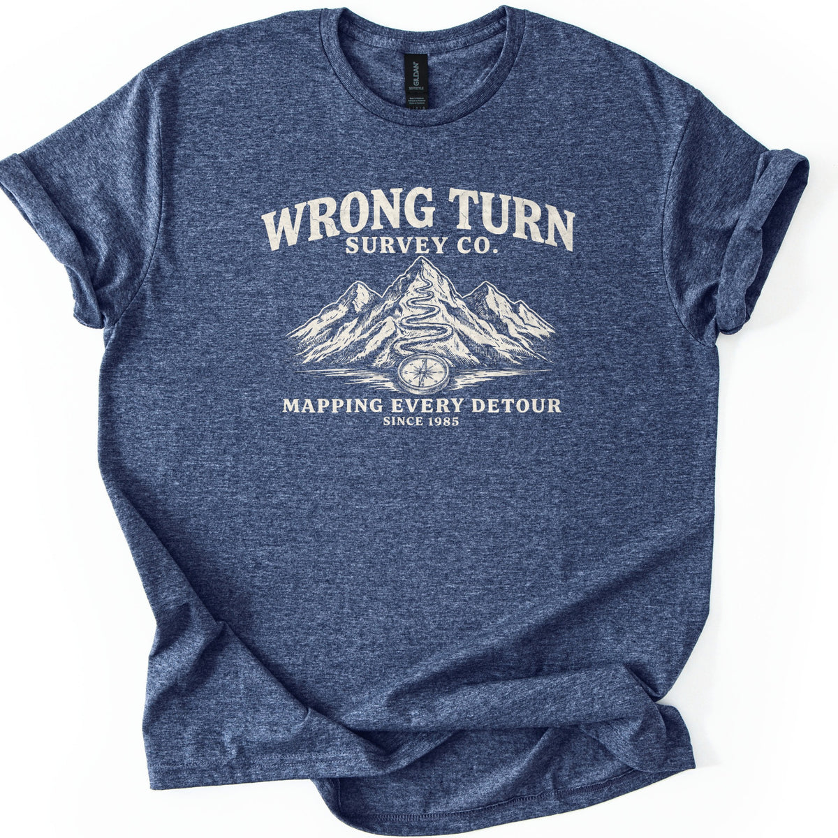 Wrong Turn Survey Co Comfort Colors T-shirt in Heather Navy, laid flat. Cream print reads Wrong Turn Survey Co. above sketched mountains with a winding trail up the tallest peak and a compass at its base, then Mapping Every Detour, Since 1985. Sleeves cuffed on a white background.