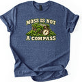 Moss Is Not a Compass Comfort Colors T-shirt in Heather Navy, laid flat. The words Moss Is Not arch above a moss-covered tree stump with a brass pocket compass leaning against it, with A Compass printed below. The shirt lies on a white background with cuffed sleeves.