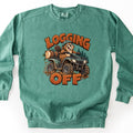 Logging Off Comfort Colors crewneck sweatshirt in Light Green, laid flat. A grinning cartoon beaver in a green helmet rides a muddy green ATV hauling a log past pine trees, between the words LOGGING OFF in orange wood-grain letters. The sweatshirt lies on a white surface.