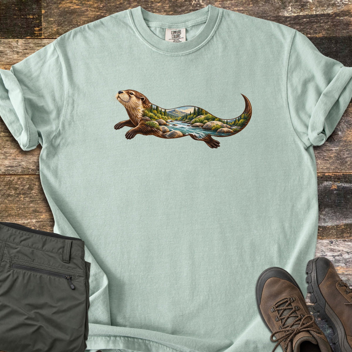 Otter River Bend Comfort Colors T-shirt in Bay, laid flat. A river otter swims across the chest, its body filled with a mountain river scene of rushing water, boulders and pine trees. Olive pants and brown hiking boots lie beside it on weathered wood planks.