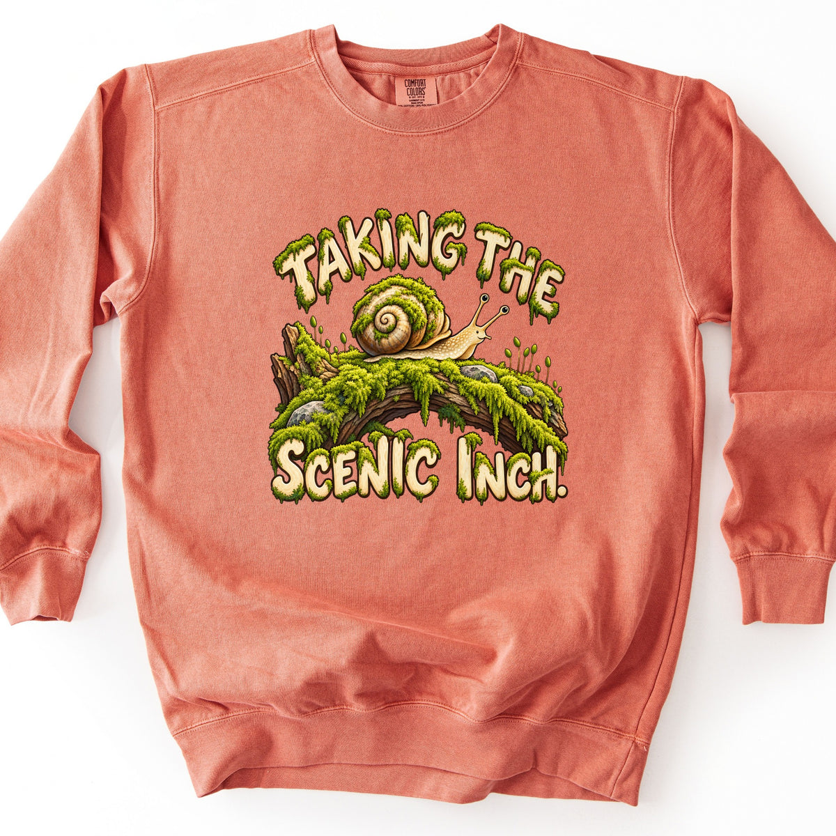 Taking the Scenic Inch Comfort Colors crewneck sweatshirt in Terracotta, laid flat. A cartoon snail with a moss-covered shell crawls along a mossy log between the words Taking the Scenic Inch. in cream letters dripping with green moss. The sweatshirt lies on a white surface.