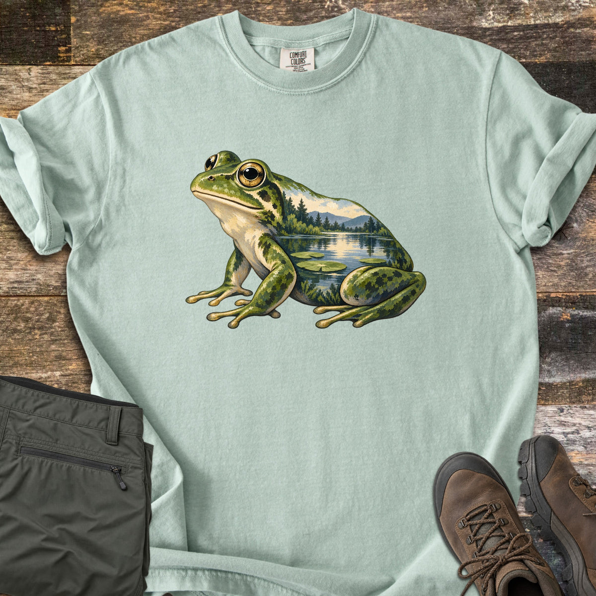 Lily Pond Frog Comfort Colors T-shirt in Bay, laid flat. A painted green frog sits in profile, its body filled with a lake scene of pine trees, mountains, and lily pads. Folded olive pants and brown hiking boots lie beside it on weathered wood boards.
