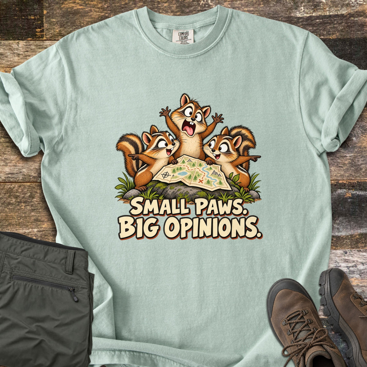 Small Paws. Big Opinions. Comfort Colors T-shirt in Bay, laid flat. Three cartoon chipmunks argue over a trail map on a mossy rock, two pointing opposite ways, above the words SMALL PAWS. BIG OPINIONS. Folded olive hiking pants and brown hiking boots sit beside it on weathered wood.