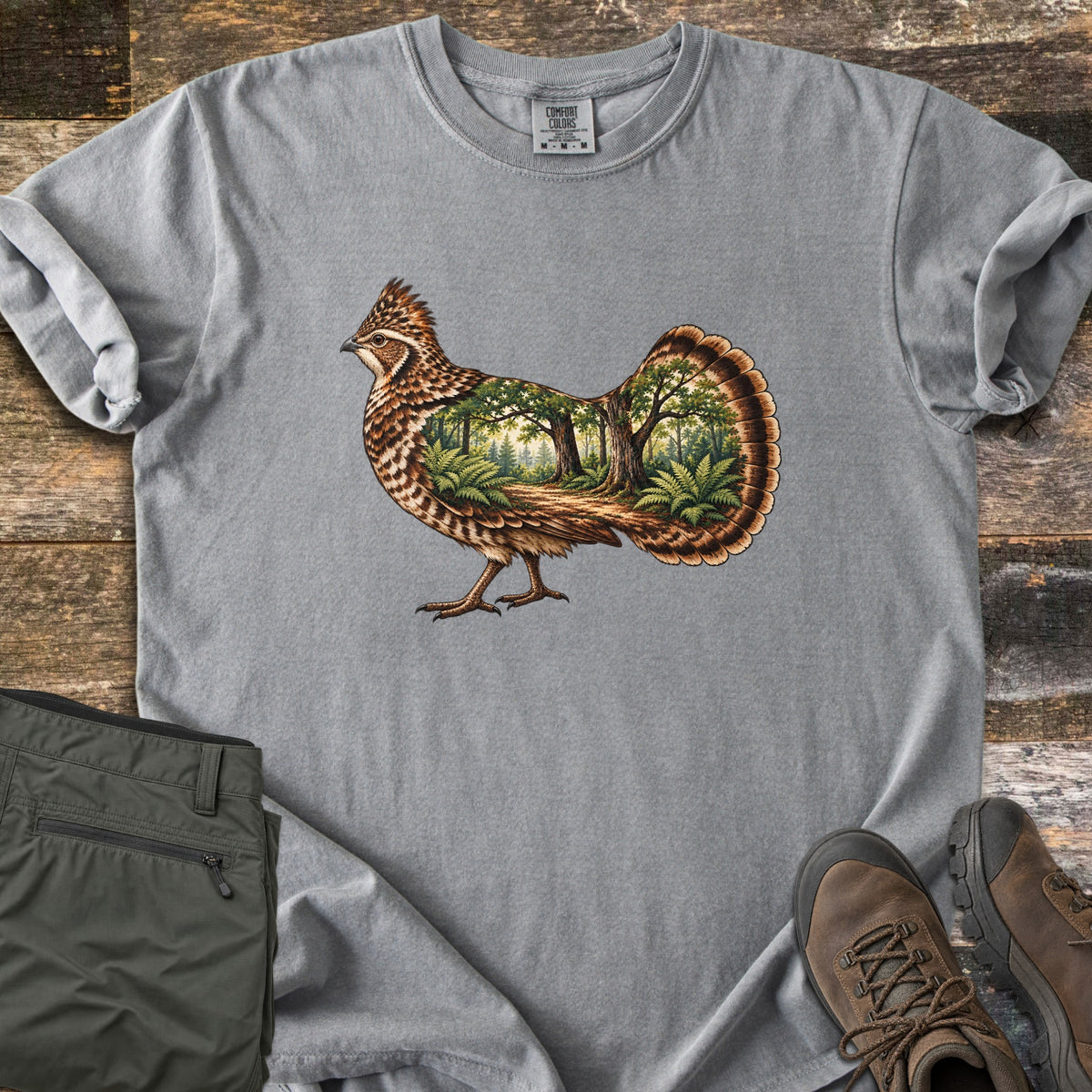 Ruffed Grouse Woods Comfort Colors T-shirt in Grey, laid flat. A detailed ruffed grouse with a fanned tail, its body filled with a forest scene of oak trees, ferns and a dirt path. Folded olive pants and brown hiking boots sit beside it on weathered wood planks.