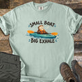 Small Boat Big Exhale Comfort Colors T-shirt in Bay, laid flat. A smiling cartoon otter in an orange life vest paddles a teal kayak between the words Small Boat Big Exhale. The shirt lies on weathered wood beside folded olive hiking pants and brown hiking boots.