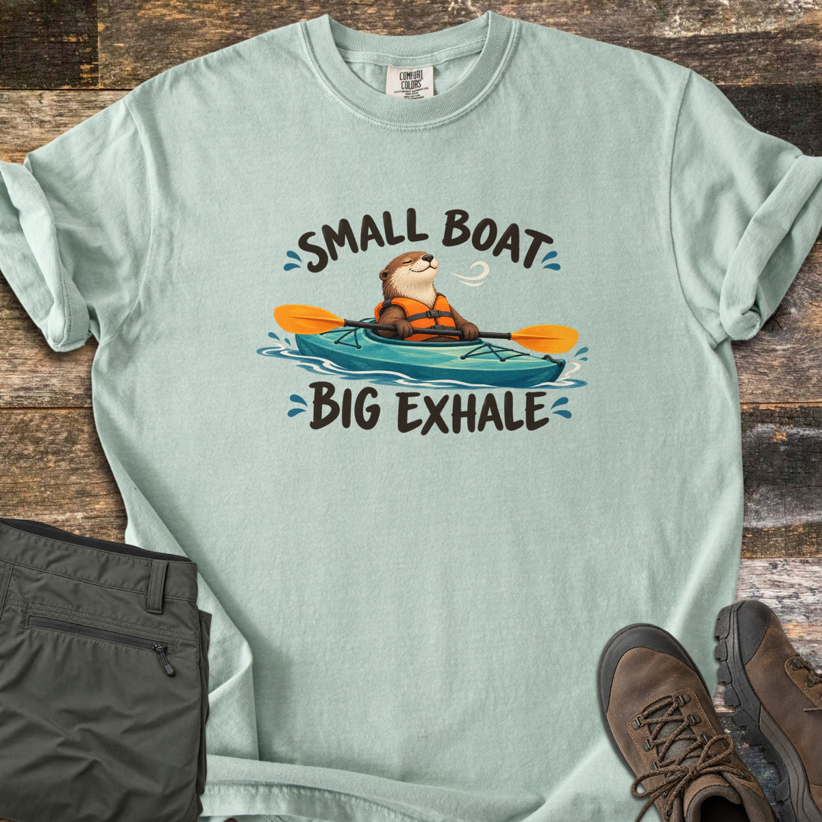 Small Boat Big Exhale Comfort Colors T-shirt in Bay, laid flat. A smiling cartoon otter in an orange life vest paddles a teal kayak between the words Small Boat Big Exhale. The shirt lies on weathered wood beside folded olive hiking pants and brown hiking boots.