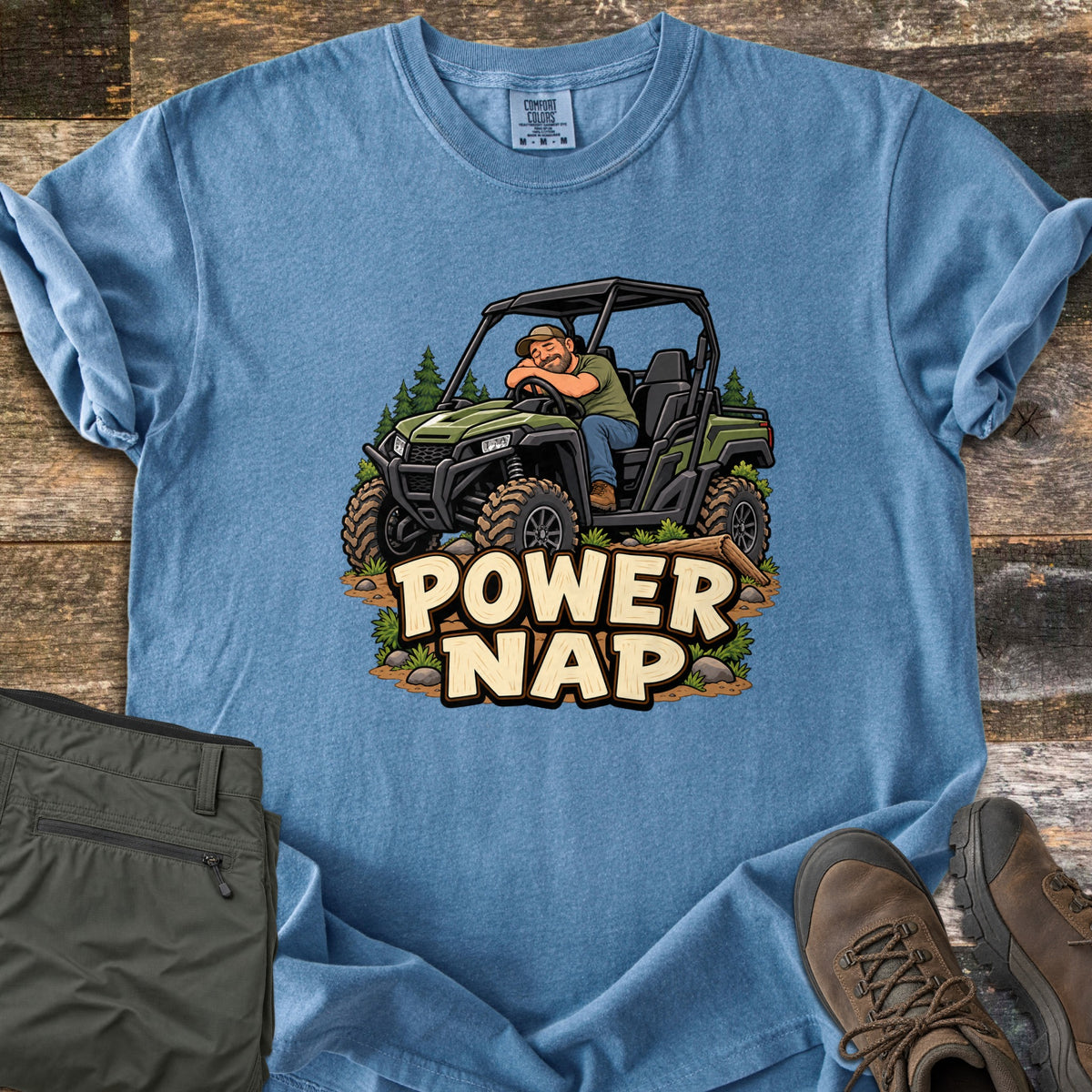 Power Nap Comfort Colors T-shirt in Blue Jean, laid flat. A cartoon man in a cap naps with his head on the steering wheel of a green side-by-side UTV among pine trees, above the words Power Nap. Folded olive pants and brown hiking boots sit on a weathered wood surface.