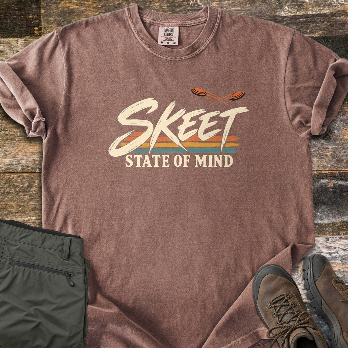 Skeet State of Mind Comfort Colors T-shirt in Espresso, laid flat. Brushy cream letters spell Skeet over orange, yellow and teal stripes, with two orange clay targets flying above and State of Mind below. Olive pants and brown hiking boots sit on rustic wood planks.