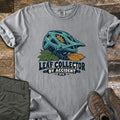 Leaf Collector By Accident Comfort Colors T-shirt in Grey, laid flat. A teal mountain bike helmet with an orange oak leaf in its vent sits on green and gold leaves above the words Leaf Collector By Accident MTB. Folded olive pants and brown hiking boots lie on weathered wood.