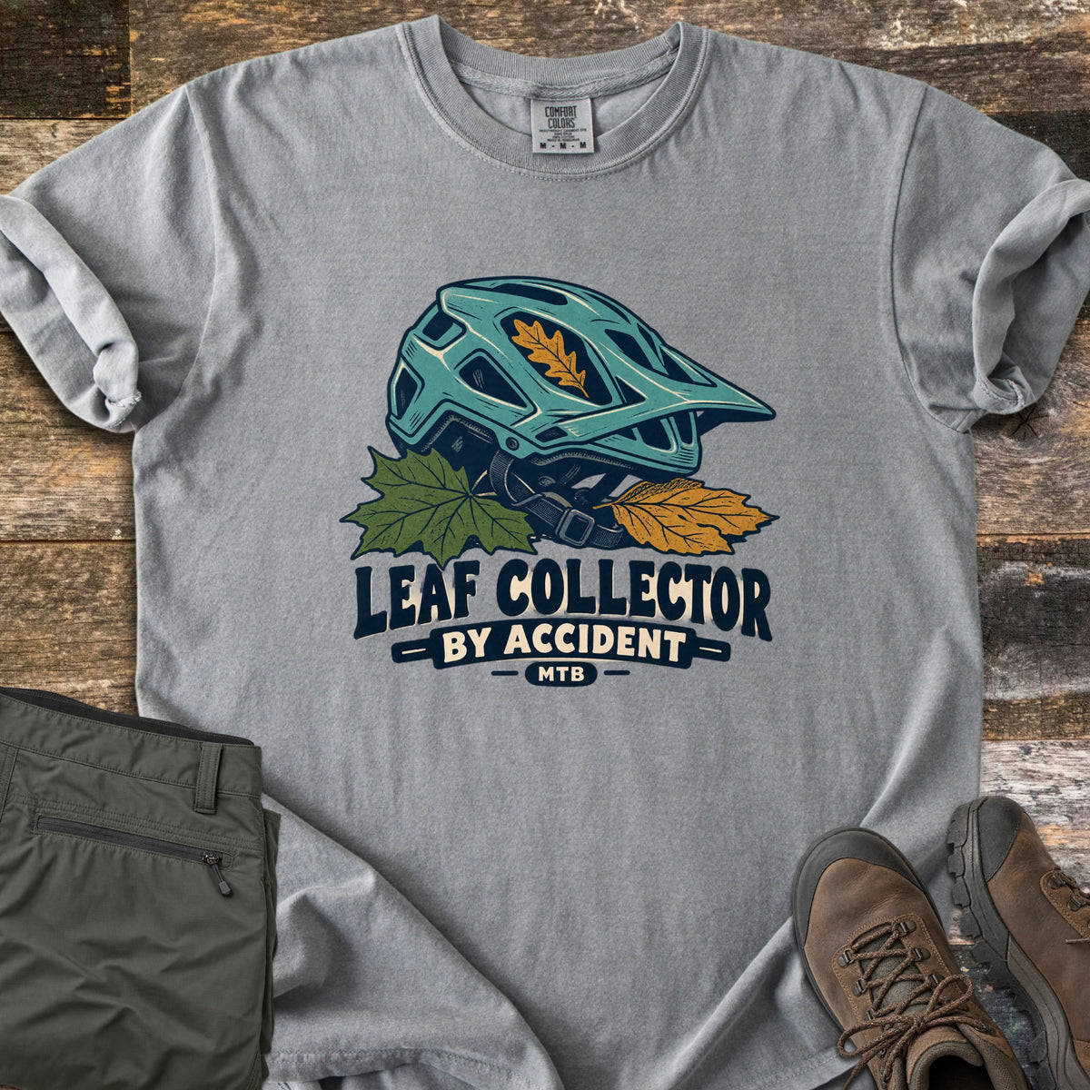 Leaf Collector By Accident Comfort Colors T-shirt in Grey, laid flat. A teal mountain bike helmet with an orange oak leaf in its vent sits on green and gold leaves above the words Leaf Collector By Accident MTB. Folded olive pants and brown hiking boots lie on weathered wood.