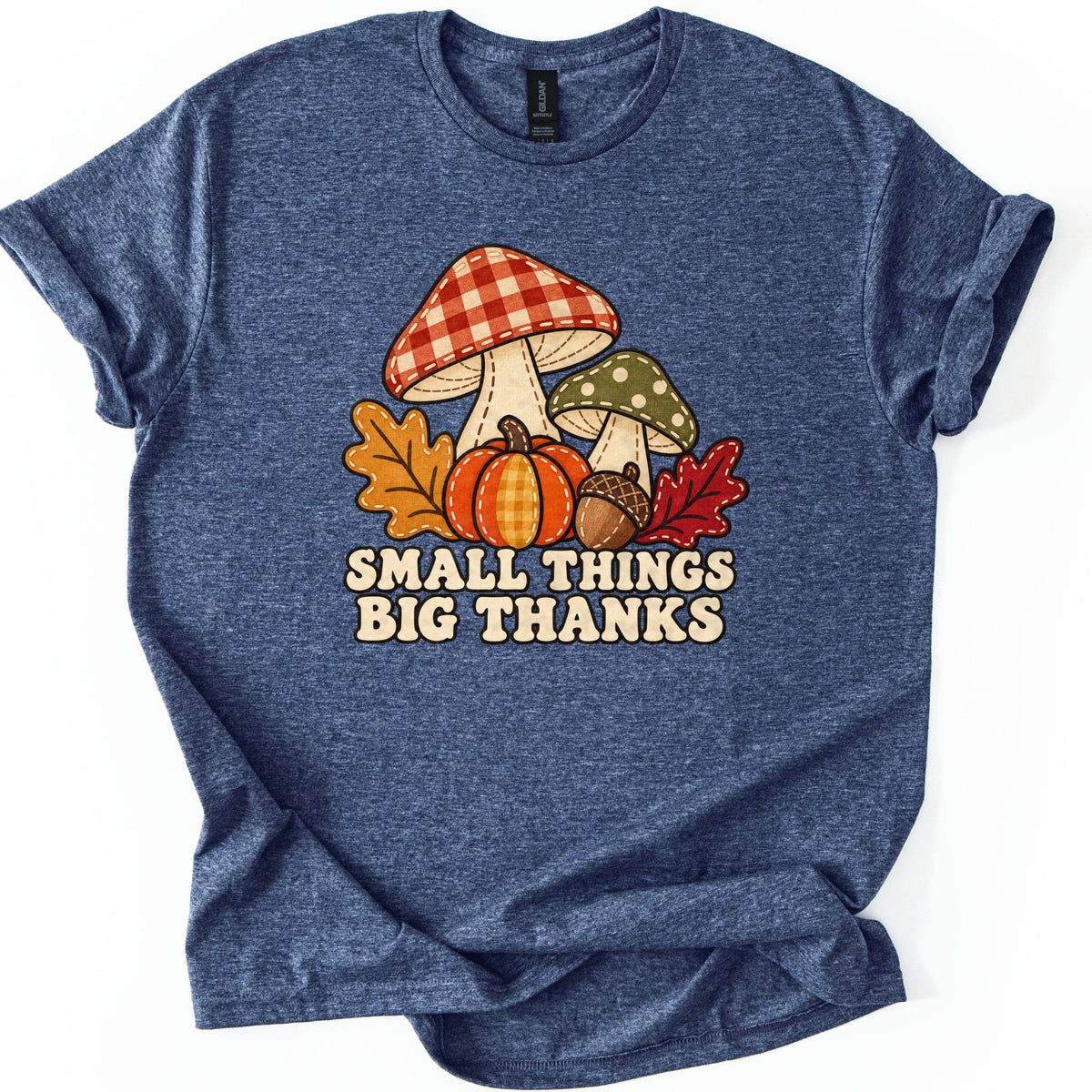 Small Things Big Thanks Comfort Colors T-shirt in Heather Navy, laid flat. A red gingham mushroom and a green polka-dot mushroom in a stitched patchwork style sit behind a pumpkin, an acorn and autumn leaves above the words Small Things Big Thanks. Sleeves rolled, on white.