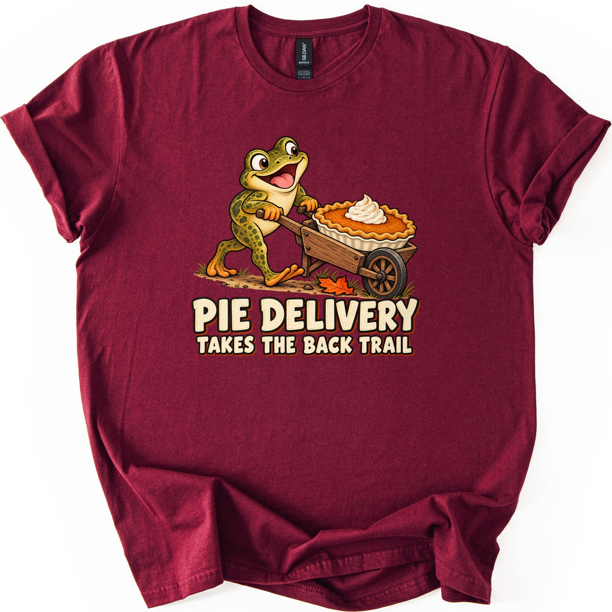 Pie Delivery Takes the Back Trail Comfort Colors T-shirt in Maroon, laid flat. A smiling cartoon frog pushes a wooden wheelbarrow holding a pumpkin pie with whipped cream along a dirt path above the words Pie Delivery Takes the Back Trail. Rolled sleeves, plain white background.