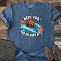 Splashy Bits Comfort Colors T-shirt in Denim, laid flat. A cartoon beaver in an orange helmet and life vest paddles a blue kayak through whitewater, framed by the words Here For The Splashy Bits. Olive pants and brown hiking boots lie beside it on weathered wood.