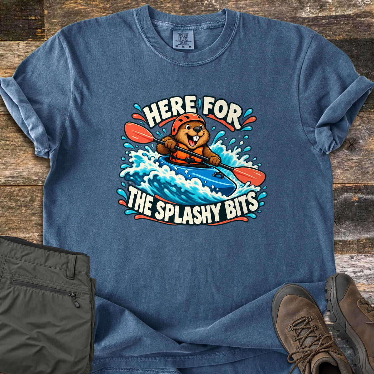 Splashy Bits Comfort Colors T-shirt in Denim, laid flat. A cartoon beaver in an orange helmet and life vest paddles a blue kayak through whitewater, framed by the words Here For The Splashy Bits. Olive pants and brown hiking boots lie beside it on weathered wood.