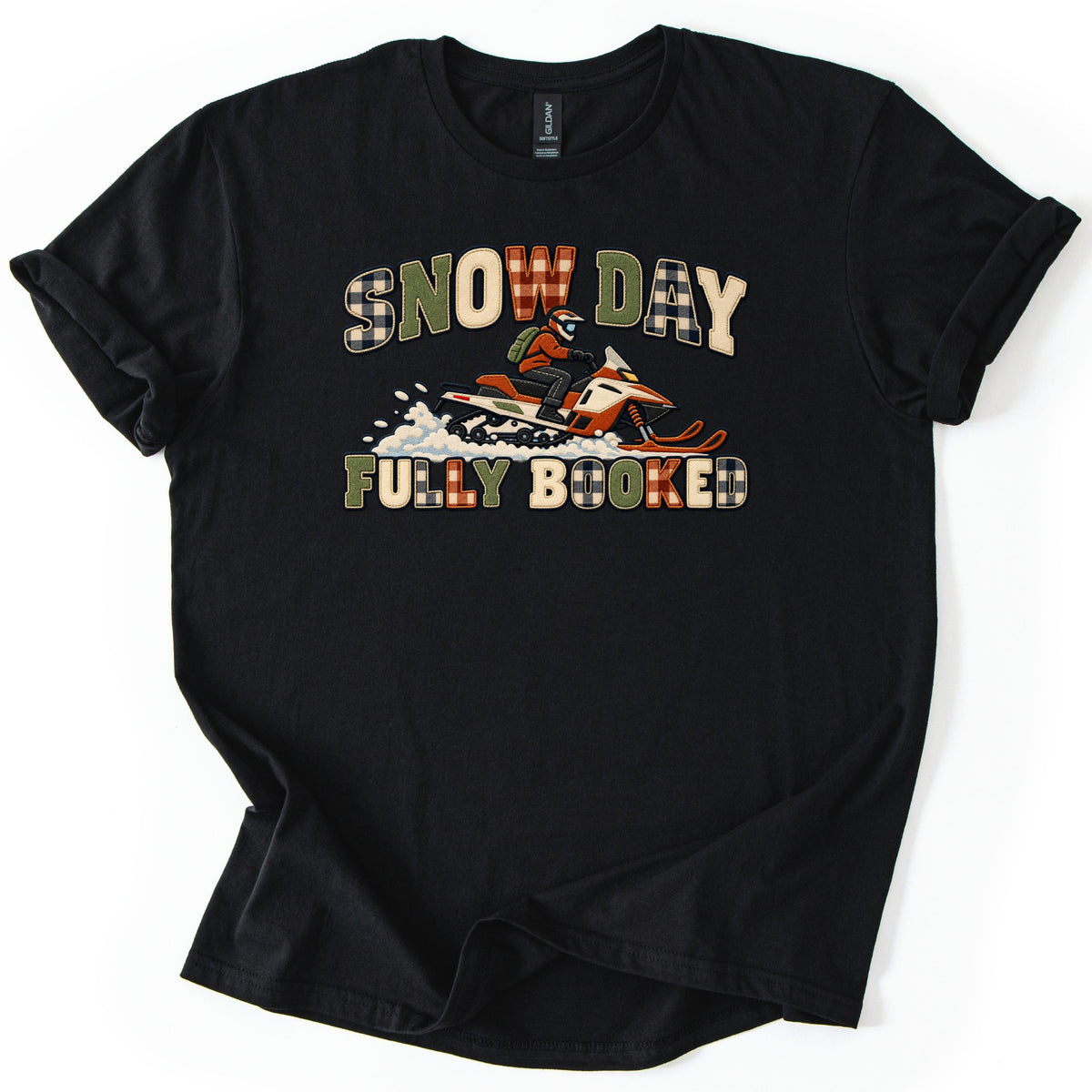 Snow Day Fully Booked Comfort Colors T-shirt in Black, laid flat. A rider in an orange jacket and helmet drives an orange snowmobile through a spray of snow, between the words Snow Day and Fully Booked in plaid, green and cream patch-style letters. The shirt lies on a white surface with rolled sleeves.