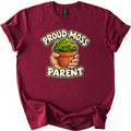 Proud Moss Parent Comfort Colors T-shirt in Maroon, laid flat. A cartoon hand holds a terracotta pot of smiling green moss between the words Proud Moss Parent in cream bubble letters. The shirt has cuffed sleeves and lies on a white background.