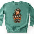 Trail Tan Comfort Colors crewneck sweatshirt in Light Green, laid flat. The front shows a mud-splattered woman in a helmet and goggles smiling in front of pine trees, with the words Trail Tan in cream and orange above an ATV kicking up mud. Shown on a white background.