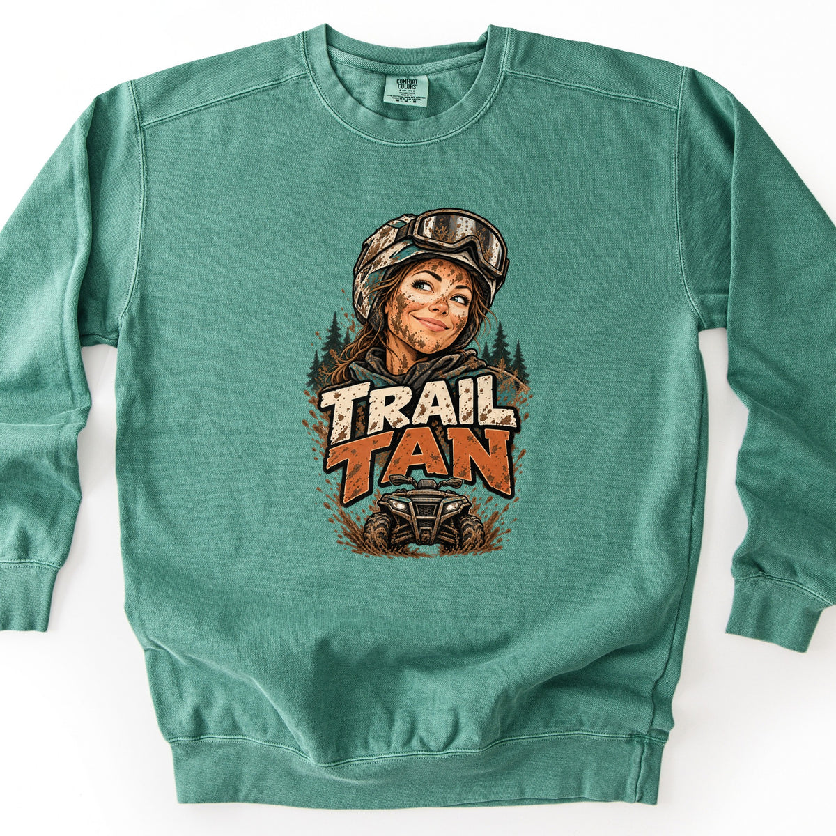 Trail Tan Comfort Colors crewneck sweatshirt in Light Green, laid flat. The front shows a mud-splattered woman in a helmet and goggles smiling in front of pine trees, with the words Trail Tan in cream and orange above an ATV kicking up mud. Shown on a white background.