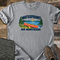 Waterfront No Mortgage Comfort Colors T-shirt in Grey, laid flat. The words Waterfront and No Mortgage frame a red canoe with a paddle on a grassy lakeshore, with pine trees, mountains and a setting sun. Green pants and brown hiking boots lie beside it on dark wood planks.