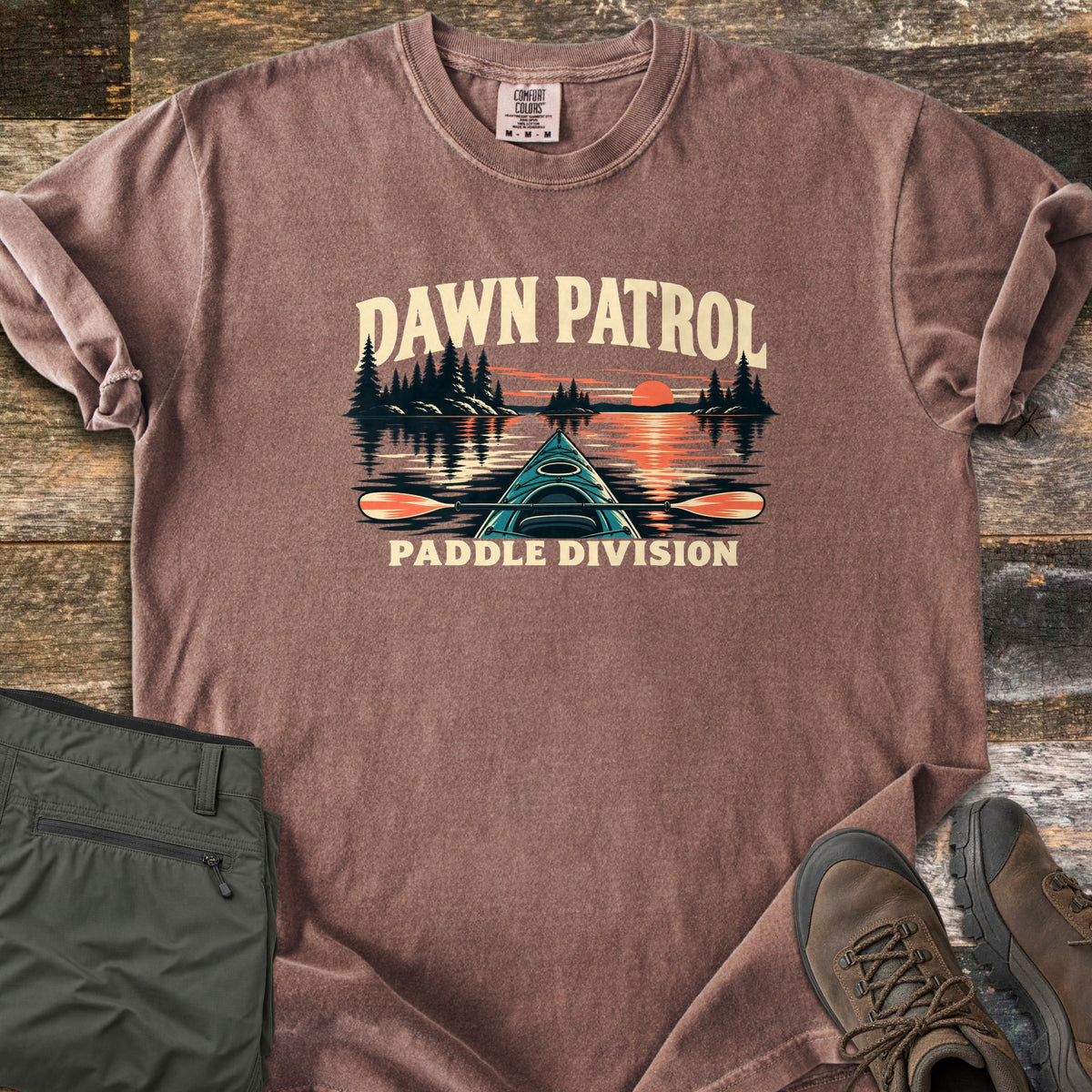 Dawn Patrol Paddle Division Comfort Colors T-shirt in Espresso, laid flat. The words Dawn Patrol arch over a teal kayak bow and paddle on a lake with pine islands and an orange sunrise, above the words Paddle Division. Olive hiking pants and brown hiking boots lie beside it on weathered wood.