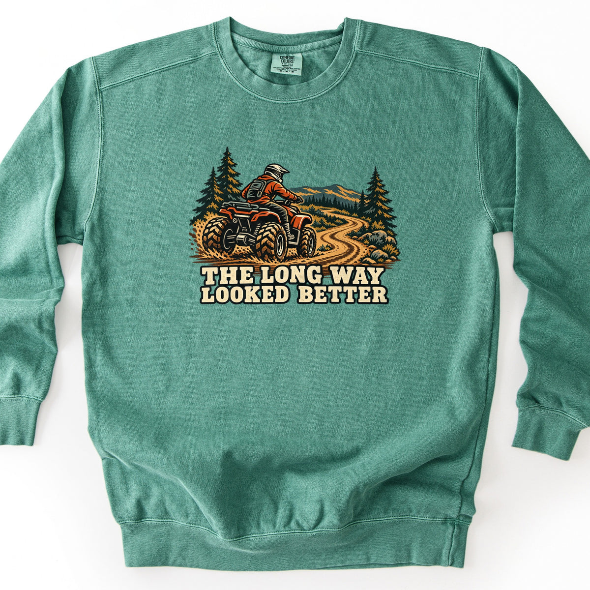The Long Way Looked Better Comfort Colors crewneck sweatshirt in Light Green, laid flat. A rider in an orange jacket and white helmet drives a red ATV up a winding dirt trail past pine trees and mountains, above the words The Long Way Looked Better. Shown on a white background.