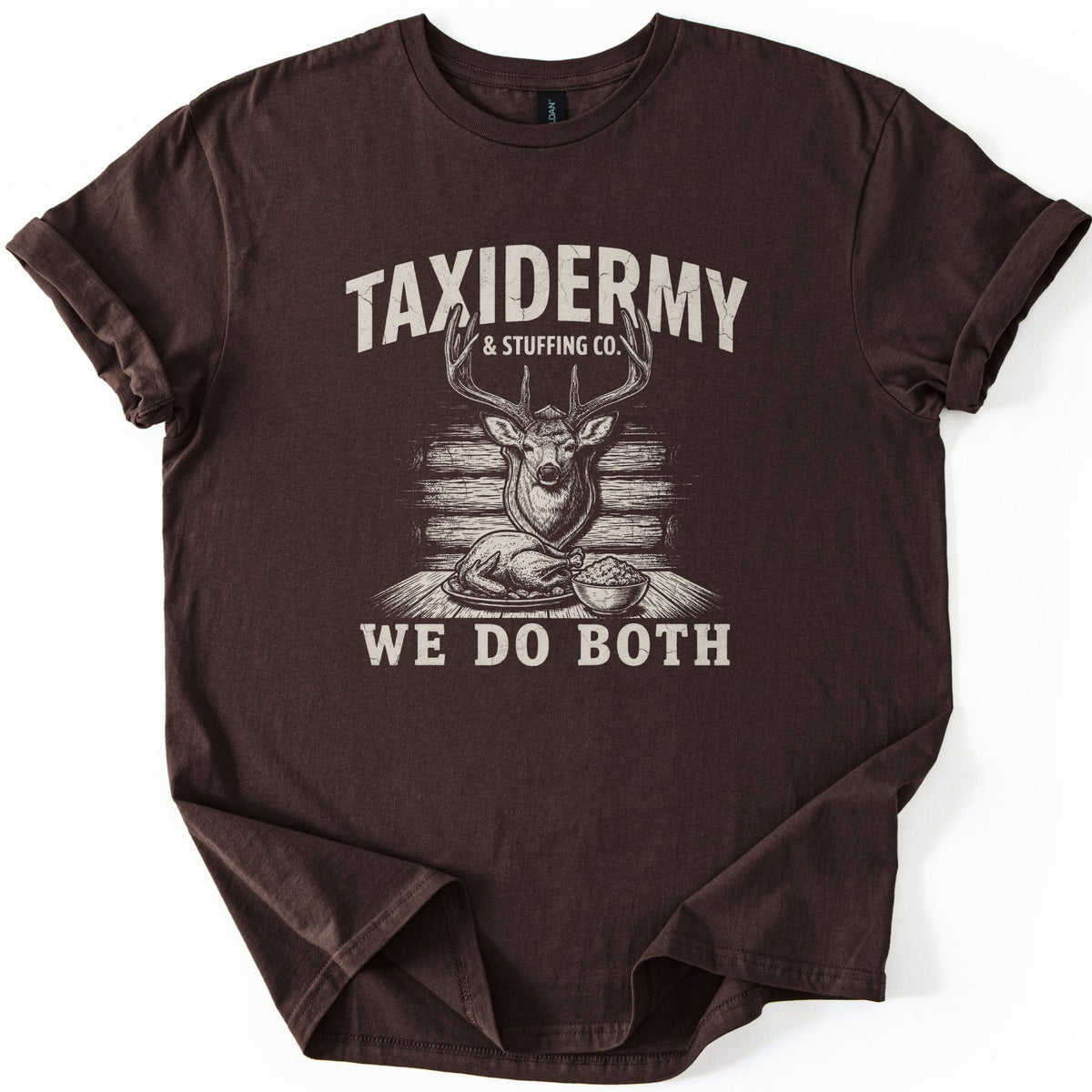 Taxidermy & Stuffing Co. We Do Both Comfort Colors T-shirt in Dark Chocolate, laid flat. A cream sketch of a mounted deer head on a log wall above a roast turkey and a bowl of stuffing, under the words Taxidermy & Stuffing Co. and above We Do Both. Sleeves are cuffed on a white backdrop.