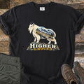 Higher Ground Comfort Colors T-shirt in Black, laid flat. A white mountain goat filled with a scene of snowy peaks, pines and a winding trail stands on rocks above the words Higher Ground. Olive hiking pants and brown boots sit beside it on weathered wood.