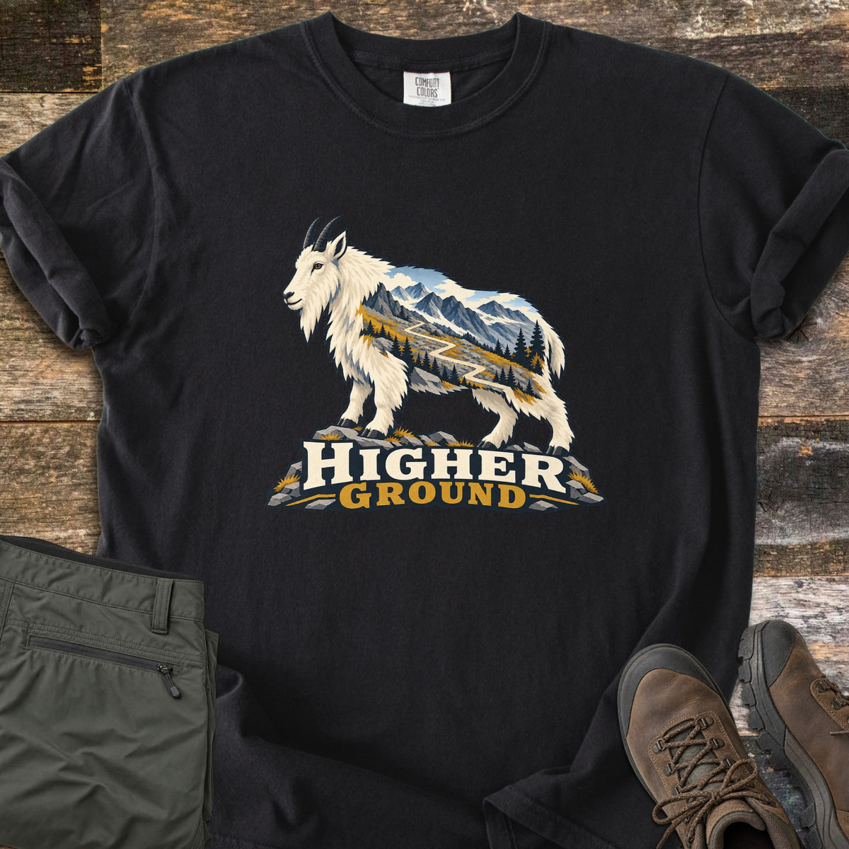 Higher Ground Comfort Colors T-shirt in Black, laid flat. A white mountain goat filled with a scene of snowy peaks, pines and a winding trail stands on rocks above the words Higher Ground. Olive hiking pants and brown boots sit beside it on weathered wood.