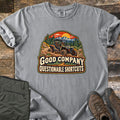Good Company Questionable Shortcuts Comfort Colors T-shirt in Grey, laid flat. An embroidered-style patch shows two riders in a side-by-side off-road vehicle kicking up dirt below mountains, pines and an orange sunset, above the words Good Company Questionable Shortcuts. Olive pants and brown boots sit on a wood floor.