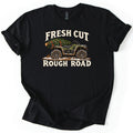 Fresh Cut Rough Road Comfort Colors T-shirt in Black, laid flat. A muddy green camo ATV with a cut evergreen tree strapped to its rear rack sits on snowy dirt between the cream words Fresh Cut and Rough Road. The sleeves are cuffed, on a white background.