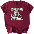 Footprints Optional Comfort Colors T-shirt in Maroon, laid flat. A grinning white furry yeti in ski goggles rides a snowmobile through spraying snow between the snow-capped words Footprints Optional. The shirt has rolled sleeves on a white background.