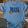Skeet Dad Comfort Colors T-shirt in Blue Jean, laid flat. Navy text reads Skeet Dad above two orange clay targets with motion trails and an oak sprig with acorns. Sleeves are rolled on a wood floor beside folded olive cargo pants and brown hiking boots.