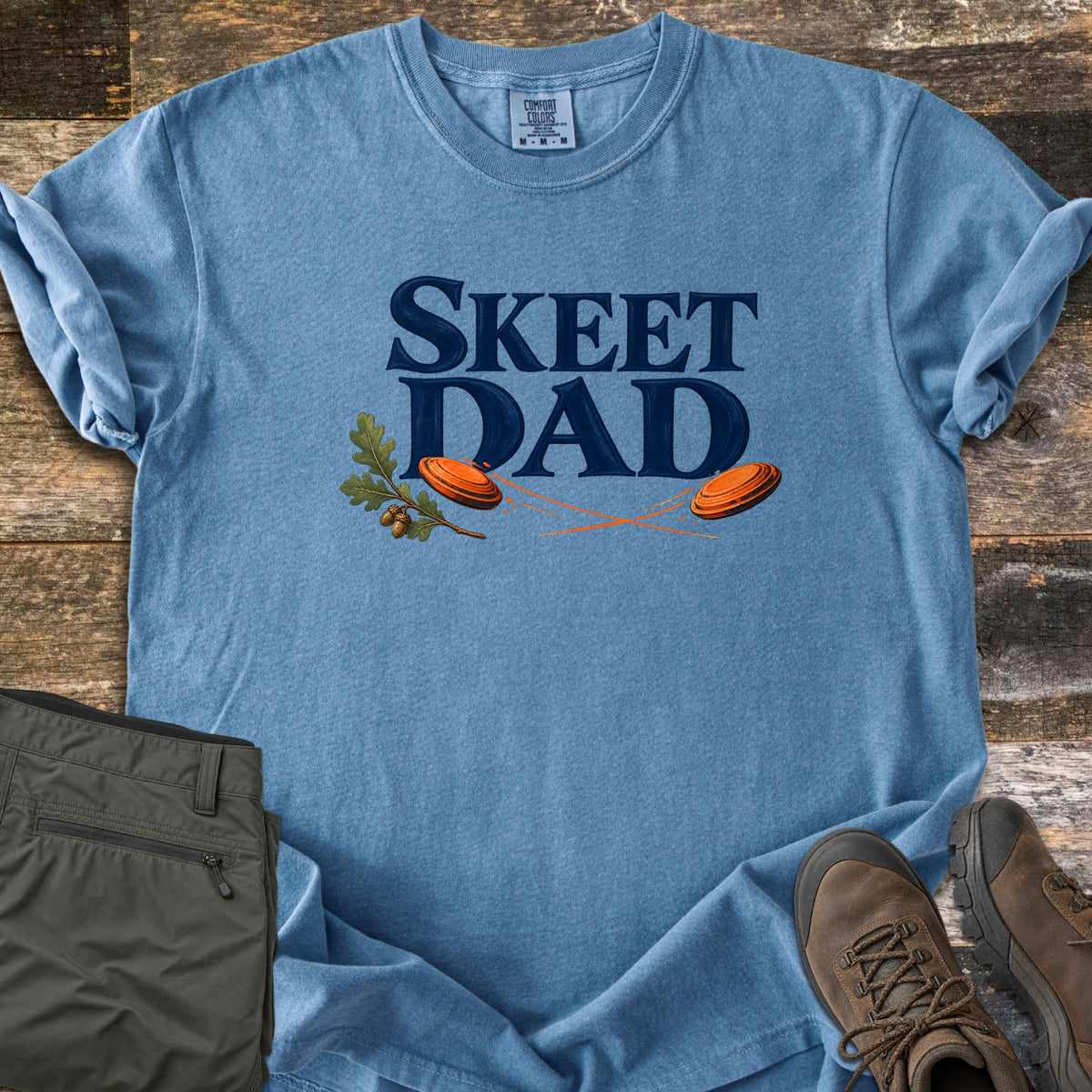 Skeet Dad Comfort Colors T-shirt in Blue Jean, laid flat. Navy text reads Skeet Dad above two orange clay targets with motion trails and an oak sprig with acorns. Sleeves are rolled on a wood floor beside folded olive cargo pants and brown hiking boots.