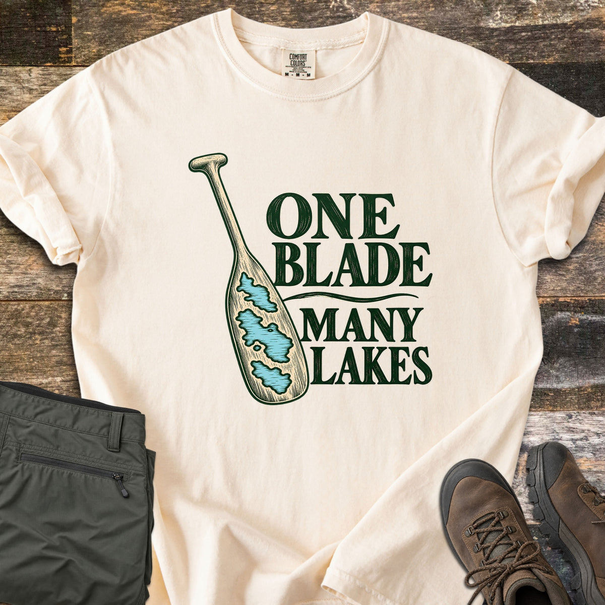 One Blade Many Lakes Comfort Colors T-shirt in Ivory, laid flat. A wooden canoe paddle with blue lakes painted on its blade sits beside the words One Blade Many Lakes in dark green. Olive hiking pants and brown hiking boots rest on weathered wood boards.