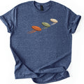 Flying Clays Comfort Colors T-shirt in Heather Navy, laid flat. Three clay targets in orange, olive green and cream fly upward in a diagonal line across the chest, each trailing motion streaks, with no words. The shirt lies on a white surface with cuffed sleeves.