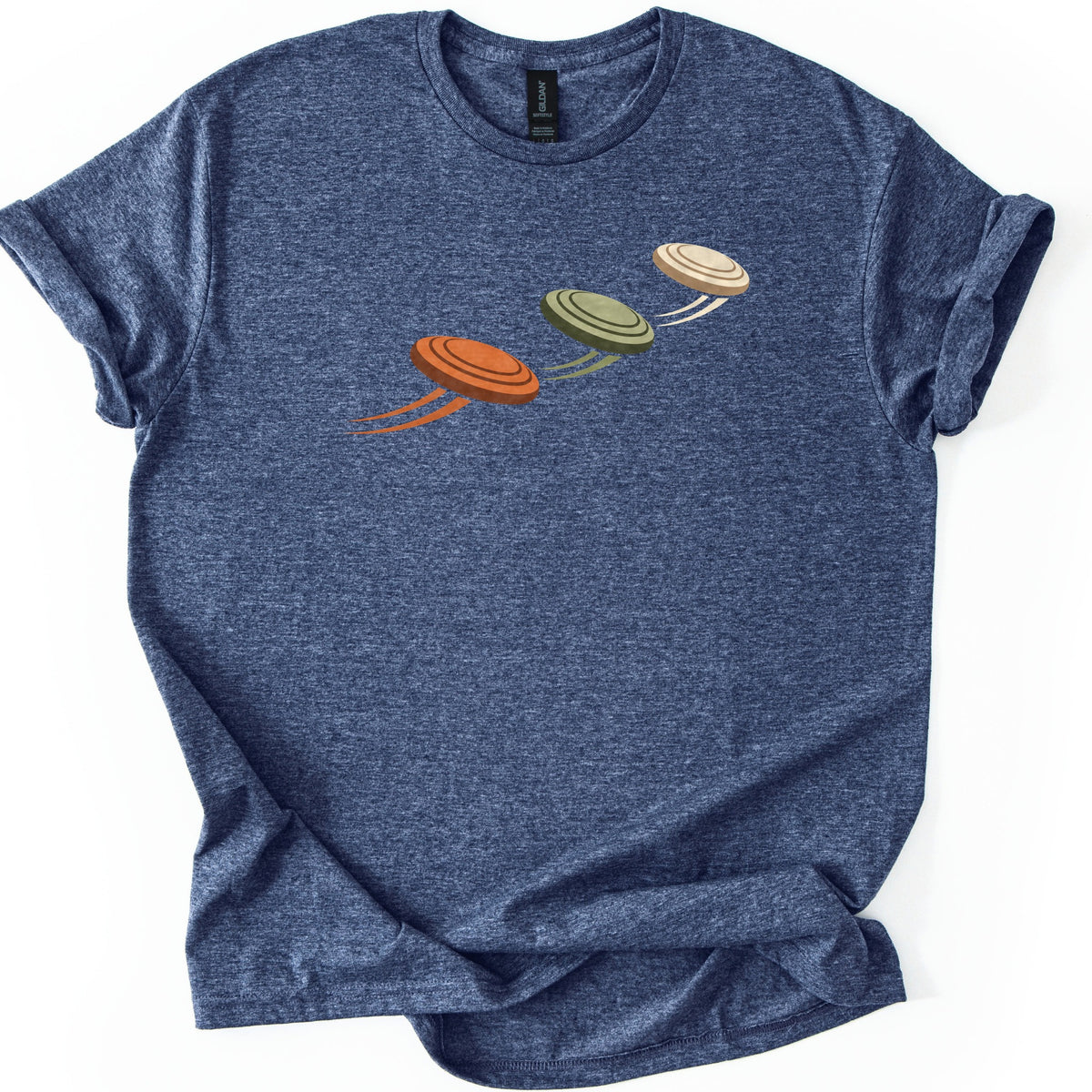 Flying Clays Comfort Colors T-shirt in Heather Navy, laid flat. Three clay targets in orange, olive green and cream fly upward in a diagonal line across the chest, each trailing motion streaks, with no words. The shirt lies on a white surface with cuffed sleeves.