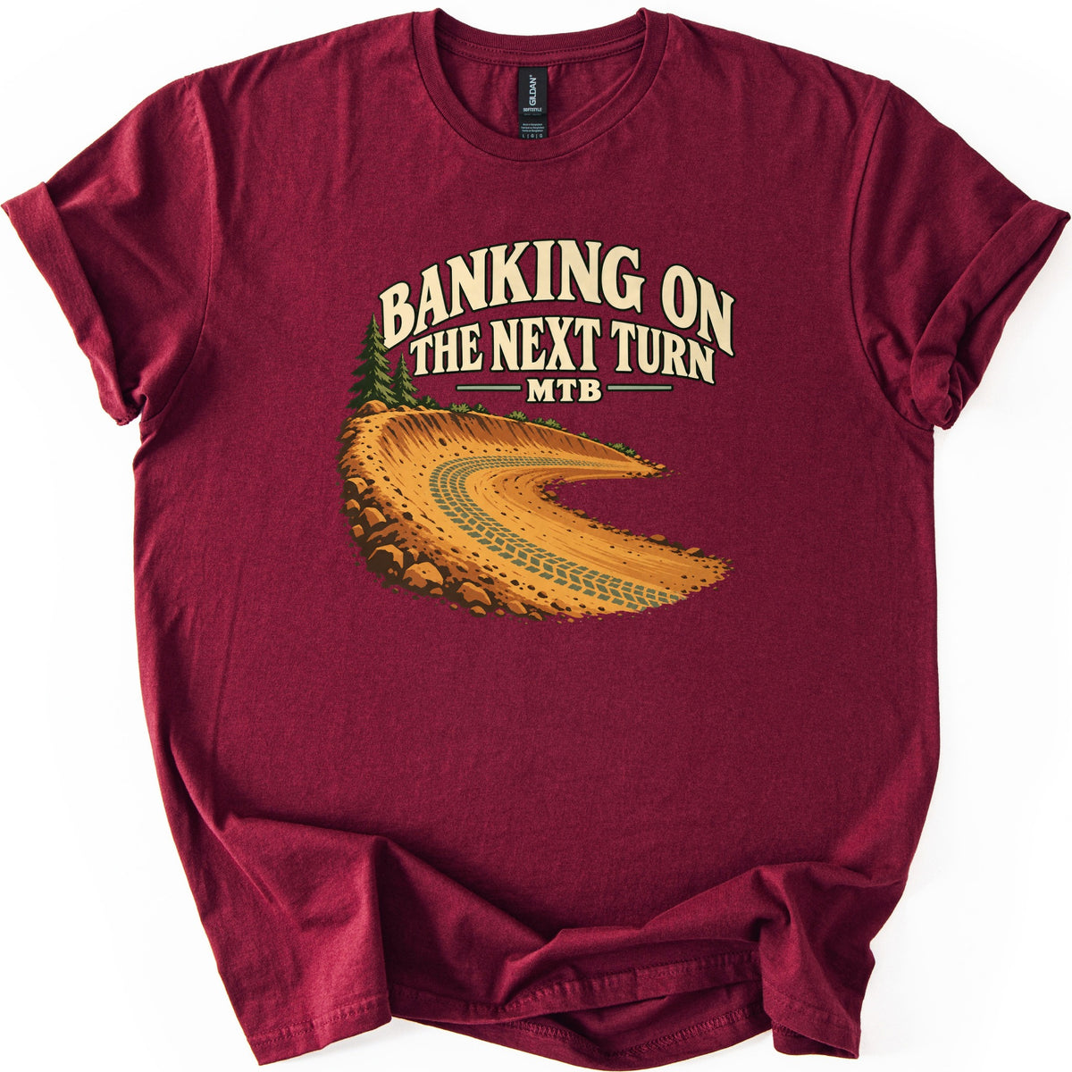 Banking on the Next Turn Comfort Colors T-shirt in Maroon, laid flat. Cream arched text reads Banking on the Next Turn, with MTB below, over an illustration of a banked orange dirt trail corner with tire tracks, rocks and pine trees. Cuffed sleeves on a white background.
