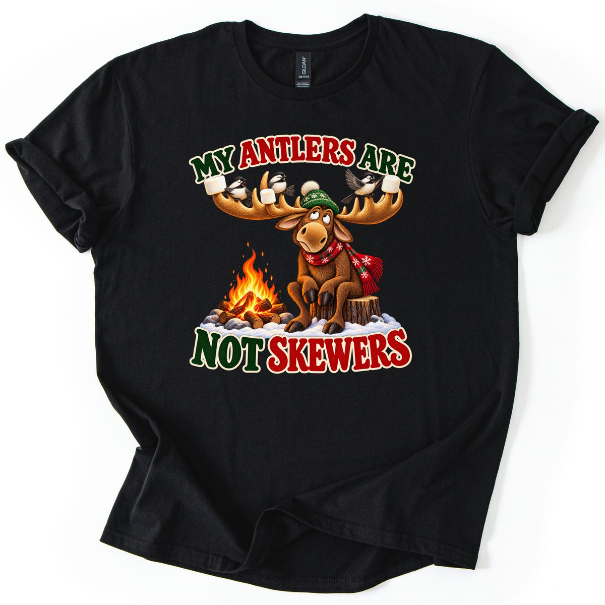 My Antlers Are Not Skewers Comfort Colors T-shirt in Black, laid flat. A cartoon moose in a green knit hat and red snowflake scarf sits on a stump by a campfire, marshmallows and chickadees on its antlers, between the words My Antlers Are Not Skewers. Shown on a white background.