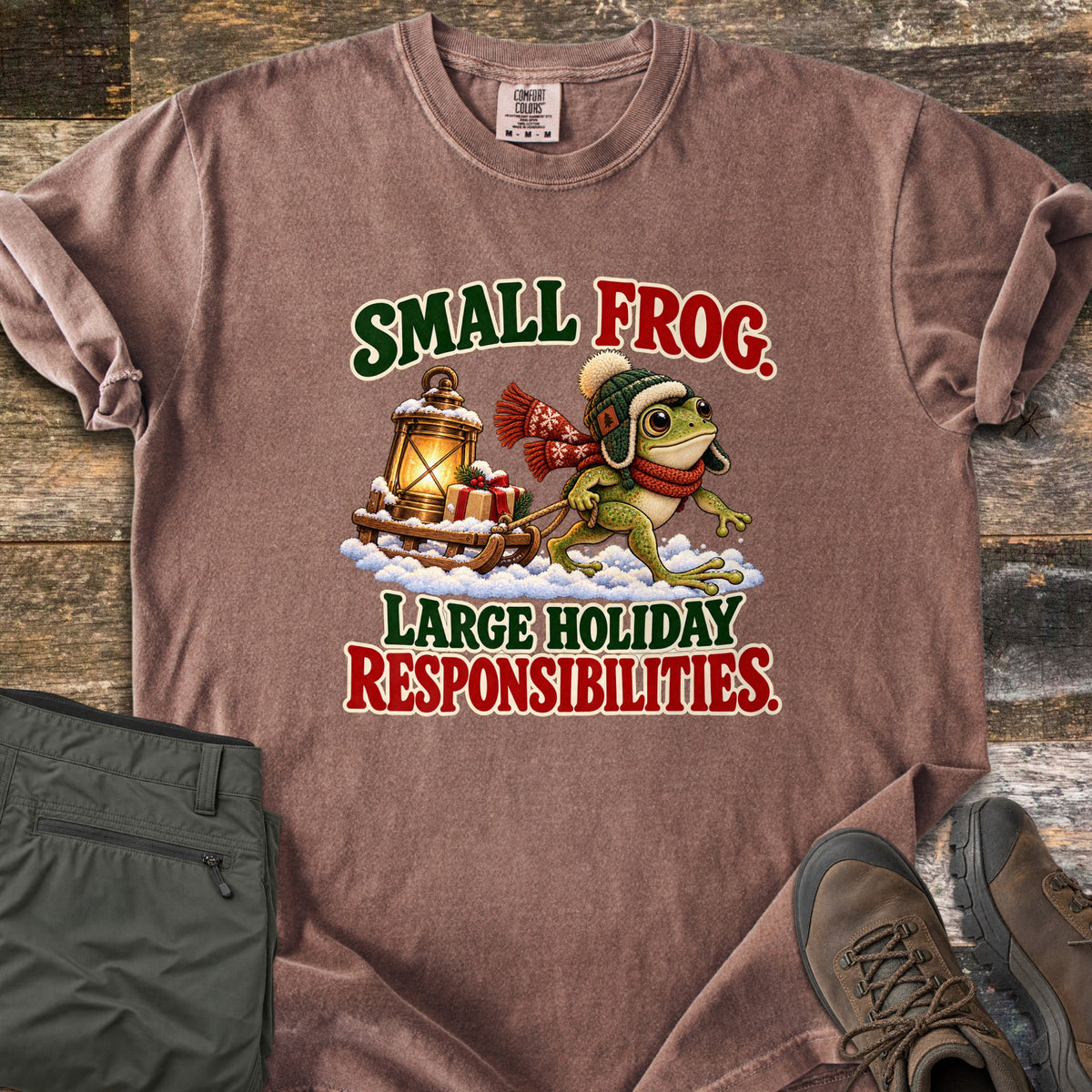 Small Frog. Large Holiday Responsibilities Comfort Colors T-shirt in Espresso, laid flat. A frog in a knit trapper hat and red scarf pulls a snowy wooden sled with a glowing lantern and gift, between the words Small Frog. Large Holiday Responsibilities. Olive pants and brown boots lie on wood planks.