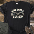 One More Loop Comfort Colors T-shirt in Black, laid flat. A cream infinity-loop dirt trail with a rider on a four-wheeler and pine trees, between the words One More Loop. Olive cargo pants and brown hiking boots lie beside it on weathered wood boards.