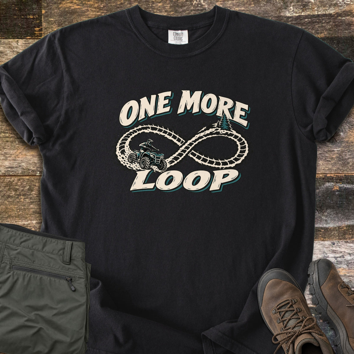 One More Loop Comfort Colors T-shirt in Black, laid flat. A cream infinity-loop dirt trail with a rider on a four-wheeler and pine trees, between the words One More Loop. Olive cargo pants and brown hiking boots lie beside it on weathered wood boards.